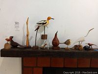 Wide view of 10 assorted wooden bird figurines arranged on wooden beam with some mounted on wood or acrylic stands.