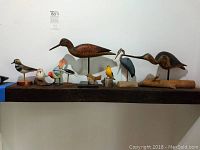 Nine assorted wooden decorative bird figurines arranged on a shelf, showing a variety of bird types and sizes.