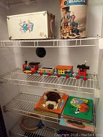 Shelf with vintage Fisher Price wooden train set of four colorful cars including engine, two middle cars, and caboose. Also visible is a metal suitcase decorated with cartoon pictures and a Playskool colored wooden blocks container.