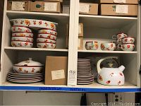 Wide view showing shelves holding stacked matching ceramic plates, bowls, cups, a teapot, and a lidded serving dish, all with multicolored floral prints and red rims.