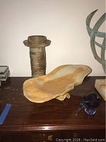Photo showing a tall textured pottery vase and a large shallow organic-shaped bowl on a wooden surface, with partial view of additional decorative objects.
