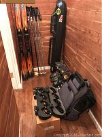 Full lot of ski equipment and accessories including skis, poles, boots, ski rack, and ski bag, arranged in corner against wooden wall.
