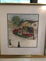 Framed watercolor painting depicting a street cafe scene with shaded tables in red umbrellas, titled 'Cafe en Arles'.
