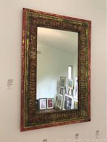 Front view of large decorative rectangular wall mirror with colorful distressed wooden frame showing red, green, and gold tones and carved linear pattern.