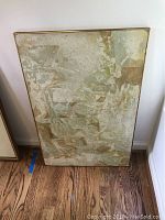 Front view of the abstract collage art with muted earth tones and textured layers leaning against a wall on a wooden floor.