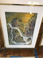 Front view of one framed watercolor painting showing turbulent water flowing through rocky cliffs with pine trees on a greenish-yellow sky background.