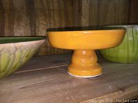 Yellow pedestal bowl shown from the side among other pottery items on a wooden shelf.