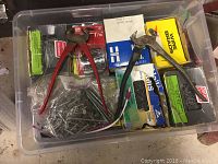 Clear plastic bin containing boxes of nails, wire brads, red-handled pliers, and loose nails.