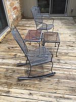 Two black metal rocking chairs with mesh seats and backs positioned on wooden deck next to two matching iron mesh side tables.