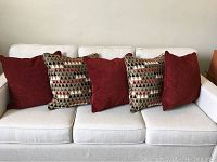 Five pillows displayed on a white beige couch. Three solid red and two larger geometric patterned pillows.