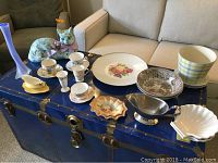 Display of various china pieces including cups, saucers, plates, a ceramic cat figurine, a tall blue vase, a silver-tone gravy boat, and a plaid planter arranged on a blue trunk.