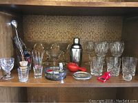 Photo showing assorted glassware including stemmed glasses, tumblers, metal cocktail shaker, metal tray, toothpick holder, cocktail picks on a wood shelf inside bookcase