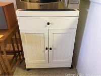 Front view of white microwave cabinet with one drawer and two paneled doors, black knobs, and caster wheels.