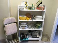 Shelf unit with various vintage and newer bakeware, kitchen utensils, folded chairs to side