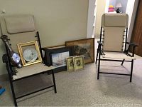 Two beige folding outdoor chairs with black frames, minor fabric marks, assorted framed artwork including floral plate, wooden clock, and prints arranged on floor and chairs.