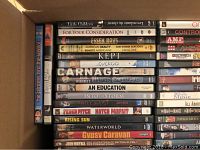 Top view of cardboard box packed with assorted pre-owned DVDs in their original cases, showing titles such as American Beauty, Waterworld, Gypsy Caravan, An Education, Into the Storm.