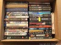 Top-down photo of multiple DVD series stacked in a cardboard box showing titles including NCIS, The West Wing, Dead Zone, The Shield, Californication, This is Wonderland, The Riches, The Mentalist, Lie to Me, My Name is Earl, Boomtown, Da Vinci's Inquest, and CSI NY series.