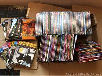 Photo showing a large assortment of DVDs, stacked vertically and horizontally in boxes and on the floor. Variety of titles and genres visible on DVD spines.