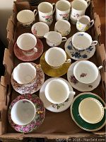 Overview of all tea cups and saucers arranged in a single layer within a cardboard box showing various floral and colored patterns.