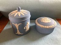 Two Wedgwood jasperware covered trinket jars, blue with white classical relief figures and decorations, displayed on a blue cloth.