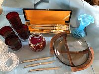 Overview of lot with ruby red glass dishes, carving set in black case with yellow lining, brown glass serving dish, blue ruffled vase, silver-colored forks, clear textured bowl.