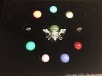 Joan Rivers Baby Bee pin with 10 interchangeable colored simulated gem backs arranged around it on black foam background.
