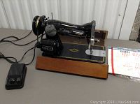 Front view of Singer sewing machine on wood base with foot pedal and manual beside it.