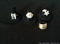 Four rings on black display stand including large rectangular stone ring, octagonal clear stone ring, floral shaped cubic zirconia ring, and wide gold-tone band ring.