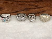 Four silver rings displayed in a row on a white surface with wood background. From left to right: silver ring with three small colored stones, wide silver ring with round and baguette stones, large round stone solitaire like ring with baguette accents, two tone ring with small stones.