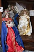 Photo showing all three dolls: two angels in white and gold robes with wings and one Madonna doll in red and blue robe holding a child doll, displayed on a wooden shelf.