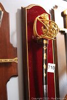 Full view showing sword mounted on red velvet and wood plaque with gold trim and ornate gold electroplated guard.