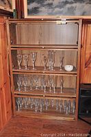 Wooden barrister-style bookcase with four shelves and glass doors. Some glassware is shown inside for display only, not included.