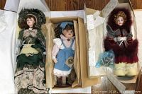 Three Boyd's Collection porcelain dolls in their boxes with paper wrapping. Left doll in green Victorian dress, center doll in blue pinafore dress, right doll in gray cloak with burgundy fur.
