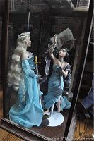 Full view of the two dolls in blue dresses inside the glass display case, showing the Lady of the Lake holding a sword and the sorceress reaching upward.
