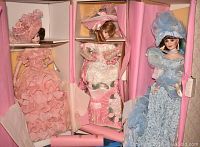 Three porcelain dolls displayed upright in open pink-lined care cases; each doll dressed in elaborate period attire with matching hats.