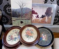 Five wall art pieces displayed together showing three oval floral prints, one print of a cow and girl, and one landscape photo with a cow.