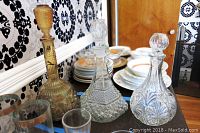 Three vintage glass decanters displayed on a dark wooden surface with plates and glasses in the background showing decorative patterns and color differentiation (amber and clear glass).