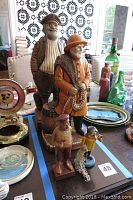 Four fisherman statues of varying sizes and materials (ceramic chalkware and wood) on a table with other collectibles and bottles in background.