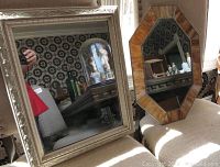 Two vintage mirrors displayed on a couch. One has a decorative white painted wood rectangular frame, the other is octagonal with a leaded slag glass frame.