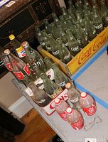 Full view of a large collection of vintage Coca-Cola and Pepsi glass bottles, including an old wooden Coca-Cola crate holding multiple bottles and a paper six-pack holder with additional bottles.