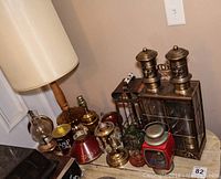 Photo showing wooden table lamp with fabric shade, various hurricane lamps and lamp oil can arranged on wooden surface.