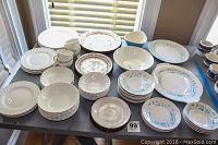 Overview of the entire ceramic dinnerware lot displayed on a black table including white, floral brown, and blue patterned plates, bowls, and cups.