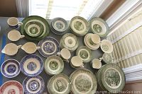 Wide shot of vintage transferware plates, cups, and creamers in green, blue and some red colors, spread on a large surface near window light.