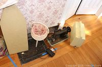 Wide shot of the sewing machine, wicker basket, and ironing board top placed on floor against wall with floral wallpaper.