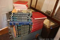 Photo showing a stack of several How-To-Do-It Encyclopedia volumes with black spines and illustrated cover images, alongside two stacks of red hardcover books, one being Practical Handyman's Encyclopedia.