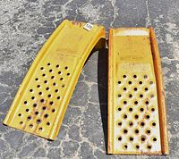 Two yellow automotive ramps side by side on asphalt, showing angle and perforated surface with rust and wear.
