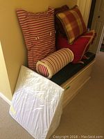 Solid wood blanket chest with several decorative pillows stacked on top, including striped and plaid patterns, along with a white bed wedge cushion leaning against the chest.