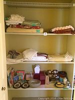 Shelf with folded bedding linens including lace trimmed blankets and sheets.