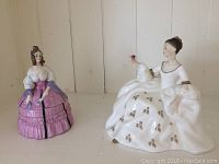 Two Royal Doulton porcelain items shown together: a figurine of a woman in a white dress with gold accents holding a small red object and a lidded box shaped as a woman in a pink dress with purple sleeves.
