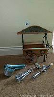 Overall view of the wood and wrought iron flower cart planter beside two vintage egg beaters and a vintage ricer on carpeted floor.